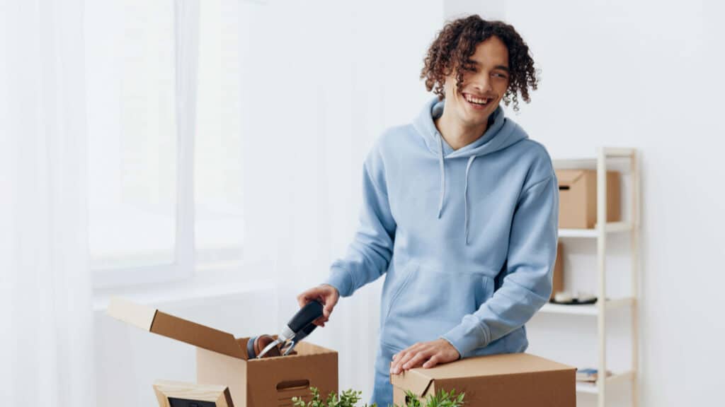 Young adult smiling while unpacking moving boxes in a bright, sunlit room; wearing a light blue hoodie and surrounded by plants and personal items.