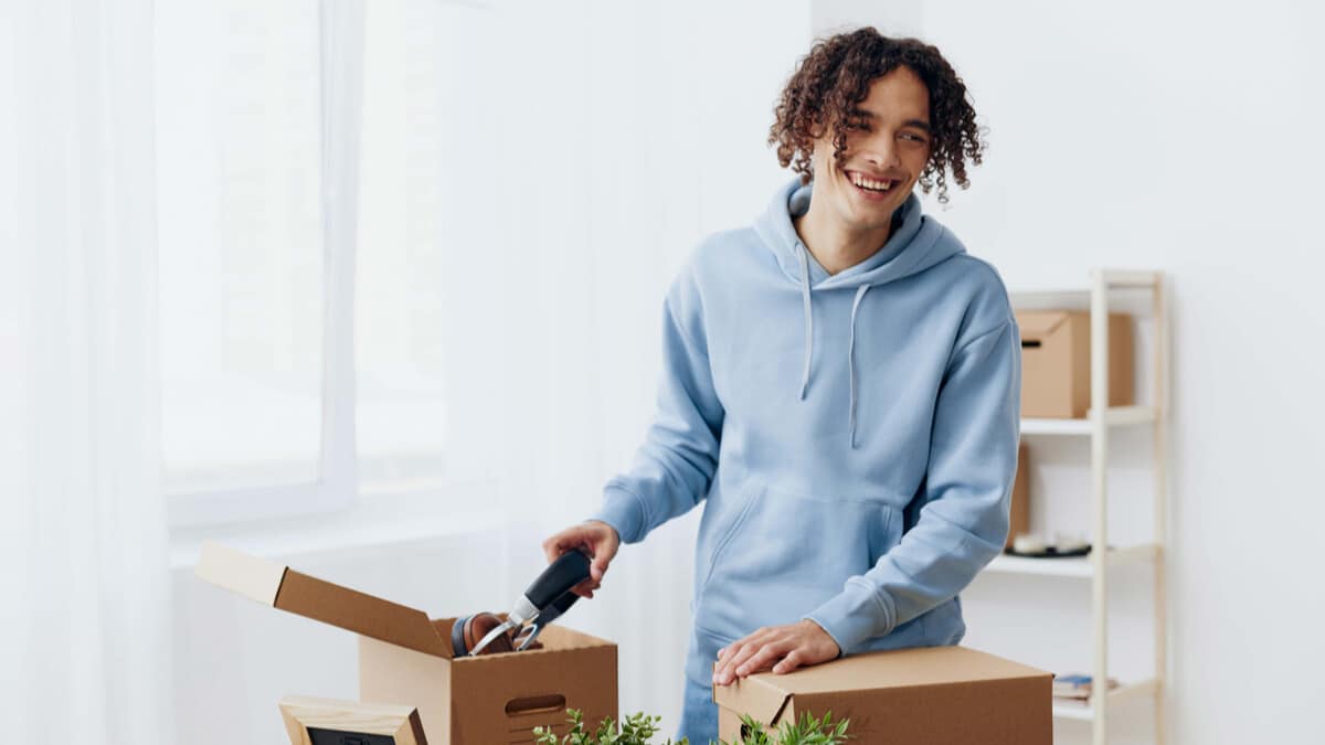 Young adult smiling while unpacking moving boxes in a bright, sunlit room; wearing a light blue hoodie and surrounded by plants and personal items.